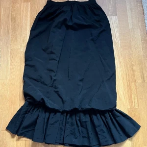 Svart midikjol med volang - Svart midikjol/maxikjol med volang nedtill. Dragkedja och knapp baktill. Köpt vintage men okänt märke, aldrig använd! toppenskick! strl S