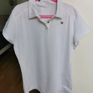 Vit träningspié från Wilson - Vit piké i funktionsmaterial för träning med fina gulddetaljer. Perfekt till bland annat tennis, padel och badminton! Knappt använd, så mycket fint skick!