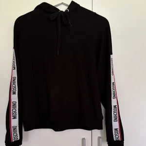 MOSCHINO HOODIE - Moschino hoodie i svart med logo på ärmarna. Något kortare i modellen. Storlek xs, passar en s också. Aldrig använd, hann tyvärr klippa bort lapparna.