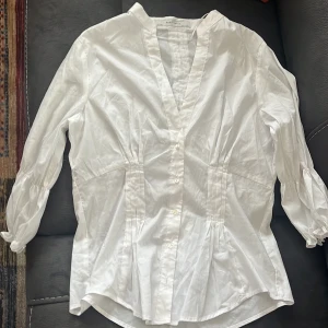 Elegant vit blus med detaljer - En vit blus med eleganta sömmar som passar för alla tillfällen då man vill lämna ett seriös intryck. Aldrig använd och tyget är av väldigt bra material.