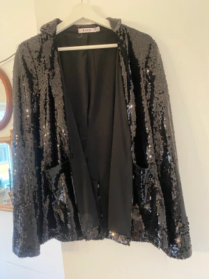 Paljettkavaj NA-KD - Svart kavaj i med paljetter.  Knappen sitter löst och måste sys på.  Annars väldigt fint skick! 