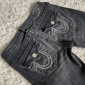 Mörkgråa True Religion jeans - Gråa True Religion jeans som är för långa. Uppvikta på bilden, använd därför inte bild som referens!! ❤️ Lågmidjade och smått bootcut/straight modell med silverdetaljer. Innerbenslängd: 84cm, midjemått: 36cm. Håll koll på min profil, säljer vintage jeans!🫶