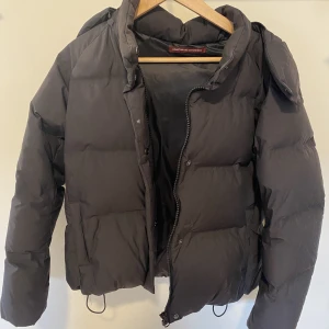 Svart bomberjacka - Vinterjacka med fodrade fickor, luva, förslutna ärmar och justerbara band vid midjan. 