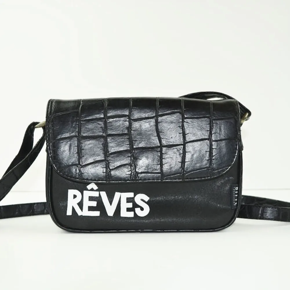 En svart basic väska med skinndetaljer och vita bokstäver nedtill. Lång axelrem som går att justera. ”Rêves” betyder  drömmar på franska. . Väskor.
