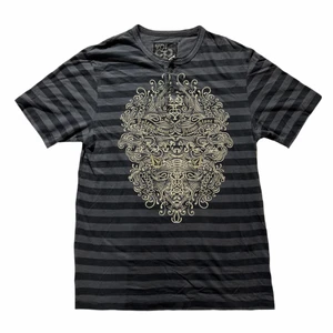 Grå randig t-shirt - Cool grå randig t-shirt med tryck på framsidan och knappar vid kragen. Tröjan har några pyttesmå hål.