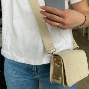 Beige Väska - Snygg beige axelremsväska! Väskan har rutigt sicksack sytt mönster fram, och guld detaljer på sidan. Den stängs med ett knapplås, men finns även en mindre ficka med dragkedja inuti. Väskan är i fint skick då den knappt är använd!Köparen står för frakten! Bara att höra av sig vid frågor!🤍🤍🤍