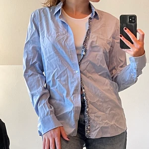 Ljusblå skjorta med blommiga detaljer - En ljusblå skjorta med blommiga detaljer på kragen och knappkanten. Passar även s&m beroende på passformen. Bra skick 💙