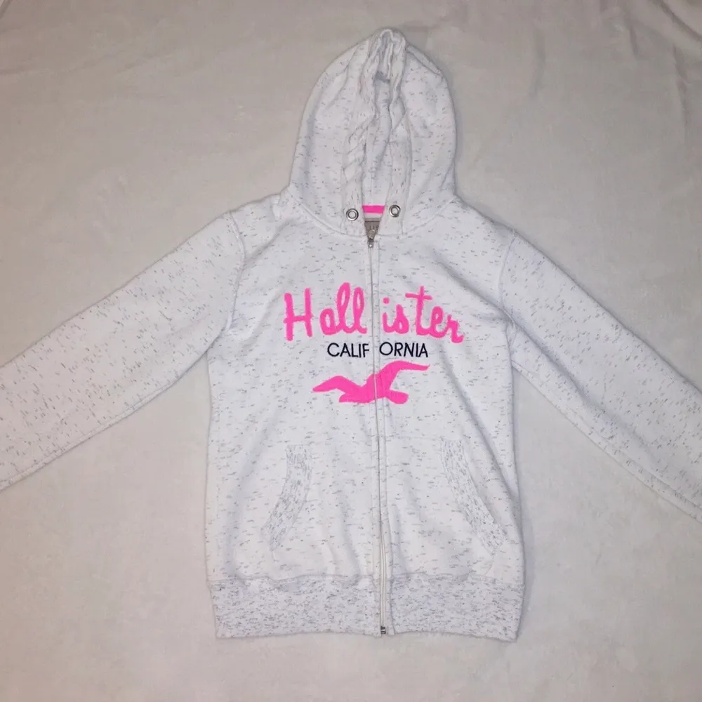 Vit-grå melerad ’hollister’ hoodie med rosa text och med dragkedja. Hoodien är en lite längre modell och är begagnad men i fint skick. På lappen står det strl XL men passar en M, då den är liten i storleken. Meddela mig vid fler frågor!😊. Hoodies.