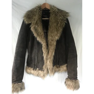 Skinn mocka mörkbrun jacka med päls st.38 - Skinn mocka mörkbrun jacka med pälsimitation st.38. Storlek 40 på lappen men liten i storlek, passar st.38 bättre. Bra skick men det finns ett märke på tyget på ärmen(kolla bilder) Två knappar. Två fickor. Längd: 68cm Material: mocka läder + pälsimitation