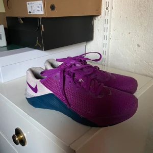 Lila Metcon skor från Nike - Säljer dessa lila metcon skor med turkosa och beiga detaljer från Nike. Jättebra tyngdlyftar/crossfit skor. De är absolut använda och lite smutsiga men funktionen är lika bra! Storleken är 37,5.  Frakt tillkommer om inget annat är överenskommet ☺️