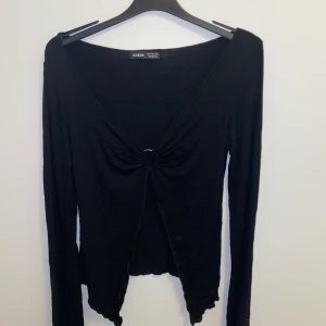 Svart Topp med fin detalj - En svart topp med en guldring i mitten som binder ihop tröjan. 