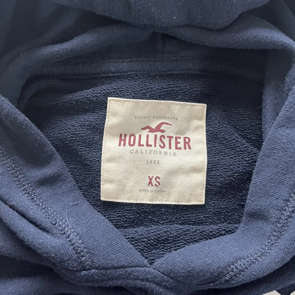 En marinblå hoodie från hollister med coola detaljer. Fint tryck på ryggen. Den är xs, men passar fint på mig som brukar ha m med bara att den är lite mer croppad då.🥰. Hoodies.