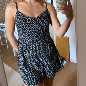 Playsuite med blommor  - Svart blommig playsuite med resor bak och knytning i sidan. Har totalt älskat denna i flera år, men skulle ändå säga att den är i väldigt fint skick! Fraktar om köparen står för frakten, möjlighet till paketpris. 