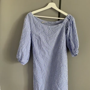 Vit/blå-randig klänning - Randig klänning med en ”one shoulder” skurning, tillhörande band till midjan. Går precis över knäna på mig som är 174cm.