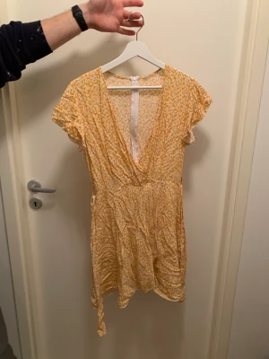 Gul småblommig klänning - Endast testad. Dragkedja i ryggen, avtagbart knytband i midjan. Tunt mjukt material. Slutar ungefär på mitten av låren.