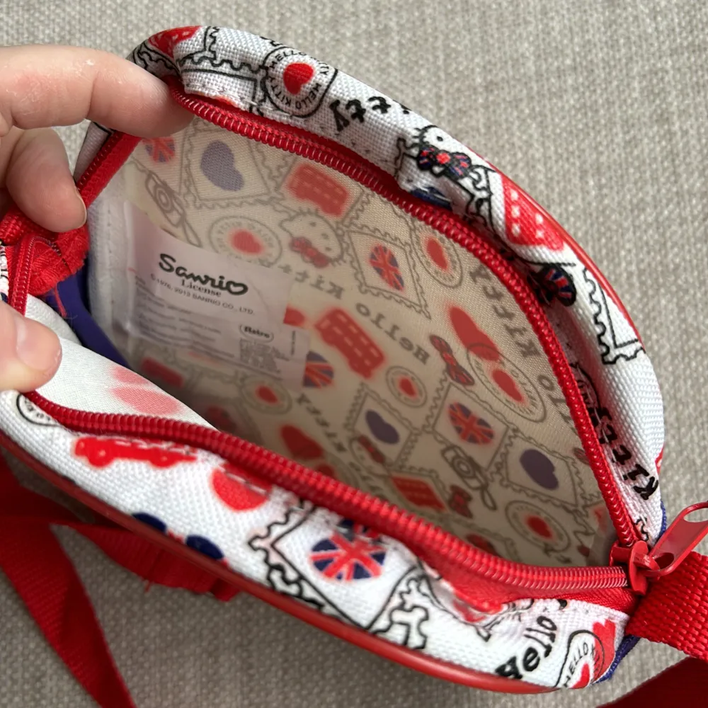 Söt liten väska med motiv av Hello Kitty Globetrotter collection -UK  Färger röd, vit och blå. Justerbar axelrem. Storlek ca 17x16 cm. Ett fack som stängs med dragkedja. . Väskor.