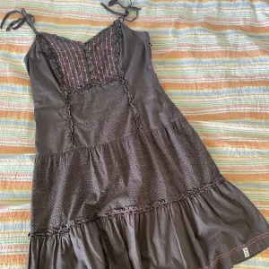 Brun klänning från de.corp - En brun klänning från de.corp med smala axelband som knyts. Klänningen har dekorativa sömmar och volanger samt en knapprad framtill. Den är tillverkad i ett lätt och bekvämt material.