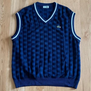 Blå stickad väst från Lacoste - En blå stickad väst från Lacoste med ett rutmönster. Västen har en V-ringning och ribbstickade kanter vid hals, ärmhål och nederkant. Lacoste-logotypen är broderad på bröstet.