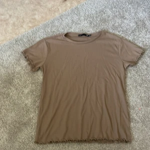 Beige t-shirt från 157 - En beige t-shirt från Lager 157. T-shirten har korta ärmar och en ribbad struktur. Den har en enkel design med vågiga kanter vid ärmslut och nederkant.