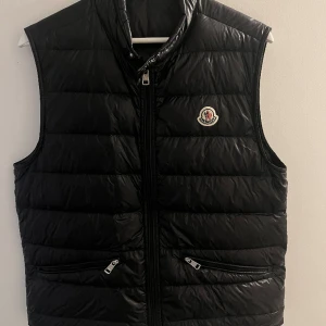 Moncler gui väst - Säljer min Moncler Gui väst då jag tänkte köpa en storlek större. Den köptes för knappt ett år sedan på Care of Carl och kvitto skickas med vid köp. Skick 7/10 då jag fick lämna in den hos en skräddare pga hål vänster bak (se bild 2)annars fin
