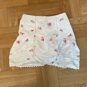 En vit kjol med rosa blommor och volanger med spets längst ned. Sitter tight runt magen och har en dragkedja vid sidan av kjolen. Jättefin passform och mönster!😍😍Slutar lite över knäna😊