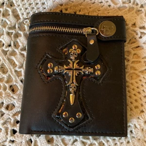 plånbok med kors o döskalle detaljer  - en plånbok som jag råkade köpa två av därför säljer jag den, är alltså helt ny! plånboken har en dragkedja framtill och flera olika fack, plats för kort och pengar 
