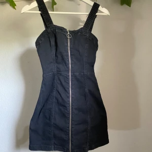 Svart hängselklänning med dragkedja  - Hängselklänning i mini- och slim fit-passform. Justerbara axelband. Aldrig använd då den inte passar längre. Köpare står för frakt 