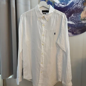 Vit långärmad skjorta från Ralph Lauren - Säljer en snygg och klassisk vit långärmad linneskjorta från Ralph Lauren. Skjortan är i bomull och har en broderad logga på bröstet. Perfekt för både vardag och fest! Den har knappar hela vägen ner och en klassisk krage. Skjortan är i nyskick och passar bra till alla tillfällen.