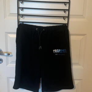 Svarta shorts från Trapstar - Säljer ett par svarta shorts från Trapstar i storlek M. De är gjorda av bomull och har en loose passform. Shortsen har en dragsko i midjan för justerbar passform och en liten logga på vänster ben. Perfekta för sommarens alla äventyr!