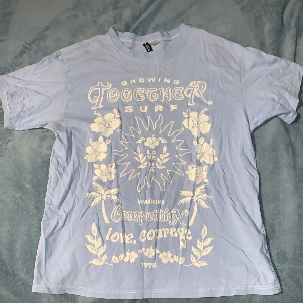 Säljer en ljusblå t-shirt med ett coolt surftryck på framsidan. Texten 'Growing Together Surf' och 'Waikiki Competition' är tryckt i vitt tillsammans med blommor och palmer. T-shirten är kortärmad och i bomull, perfekt för sommaren. Den är i bra skick och redo för nya äventyr! Oversized T-shirt.. T-shirts.