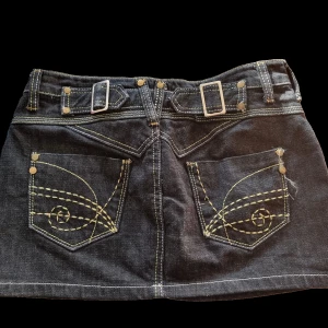 Minikjol  - Säljer en snygg svart jeanskjol med coola detaljer. Kjolen har två stora fickor bak med gul sömnad och design. Framtill finns det två fickor och metallringar. Kjolen har bälteshällor och knäpps med en knapp och dragkedja.