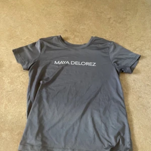 Grå t-shirt från Maya Delorez - En grå t-shirt från Maya Delorez med korta ärmar och rund halsringning. T-shirten har en enkel design med märkesnamnet tryckt på framsidan.