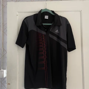Svart och grå t-shirt från Adidas - Säljer en snygg svart och grå t-shirt från Adidas i storlek XXL. Den har en cool design med röda detaljer och en krage med knappar. Perfekt för träning eller vardagsbruk. T-shirten är i bra skick och redo för en ny ägare!