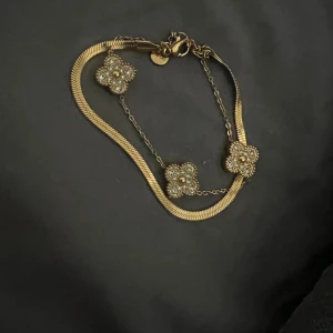 Guldarmband med pärlor och blommor - Säljer ett elegant guldarmband med tre blommor dekorerade med pärlor. Armbandet har en fin kedja och en ormlänk som ger en lyxig känsla. Perfekt för festliga tillfällen eller som en fin present.