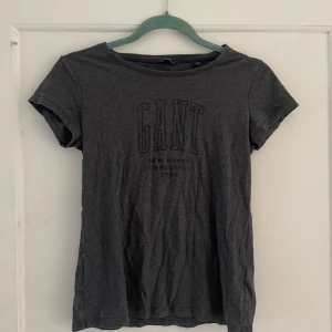 Mörkgrå t-shirt från GANT - Säljer en mörkgrå t-shirt från GANT i storlek XS. T-shirten är i bomull och har en normal passform. Den har korta ärmar och ett tryck på bröstet med texten 'GANT'. Perfekt för vardagsbruk eller avslappnade tillfällen. Köp direkt eller ge prisförslag 💛