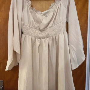 Beige klänning från Shein - Säljer en superfin beige klänning från Shein. Den har långa ärmar och en smockad midja som ger en snygg passform. Klänningen är i ett lätt och luftigt material, perfekt för sommar och vår. Den har också en söt volangdetalj vid halsen. Katt finns i hemmet, men allt jag säljer är nytvättat och rollat✨