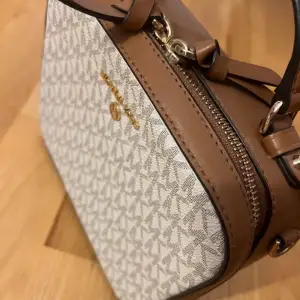 En elegant handväska från Michael Kors med ett monogrammönster i grått och vitt. Väskan har bruna detaljer och en dragkedja. Den har ett handtag för enkel bärning. Det finns även ett extra långt band som medföljer. Inköptes för:2200 Mitt pris: 850