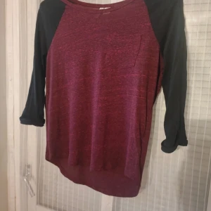 Vinröd och svart långärmad tröja - En bekväm vinröd tröja med svarta ärmar. Tröjan har en bröstficka och långa ärmar. Perfekt för höst- och vinterdagar.