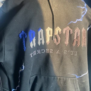 Svart hoodie från Trapstar - En svart hoodie från Trapstar med texten 'Trapstar It's a Secret' tryckt på framsidan i olika färger. Hoodien har en magficka. OBS rep