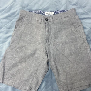 Grå shorts från Dressmann - Ett par grå shorts från Dressmann i linneblandning. Shortsen har två framfickor och två bakfickor med knappar. De har en knapp och dragkedja framtill.