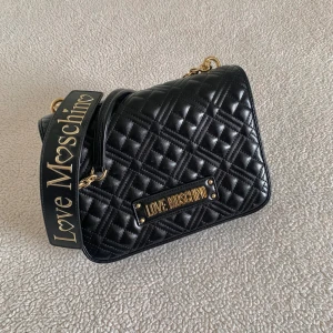 Love Moschino svart quiltad axelremsväska - Axelremsväska från Love Moschino. Endast använd i några timmar och har bara legat i dustbag och dammat sedan dess. Helt nyskick, utan anmärkningar. Kvitto från Zalando finns. Ordinariepris 2095kr. Skickas med dustbag. 