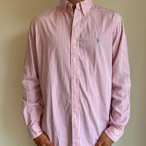 Ralph lauren  - Rosarandig Ralph Lauren skjorta i storlek L. Materialet är i lyxigt bomullssatin. Skjortan är i fint skick utan några skavanker, det finns små nästan omärkbara fläckar vid axeln. Modellen är 186 cm lång. Orginalpris ca 1500 kr och nu endast 499. 