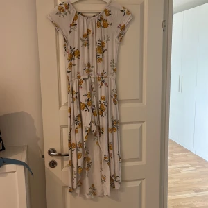 Vit blommig klänning från Lindex - En vit klänning från Lindex med ett blommigt mönster i gult och grönt. Klänningen har korta ärmar och byxor som sitter i klänningen. Den är tillverkad i Indien.