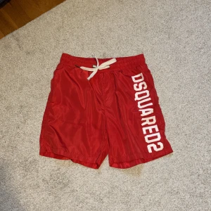 Röda shorts från Dsquared2 - Röda Dsquared2 badshorts  med vita snören och vit text på vänster ben. Shortsen har en elastisk midja och är tillverkade i ett glansigt material. Jättelite av stygnen har gått upp på ett ställe men det har inte varit några problem. 