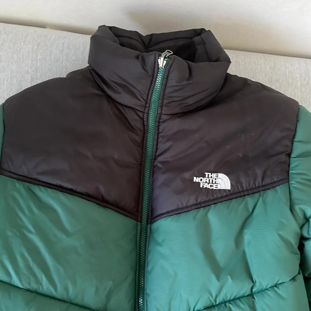 En varm och bekväm pufferjacka från The North Face i grönt och svart. Jackan har en dragkedja framtill och är quiltad för extra värmeisolering. Perfekt för kalla vinterdagar. Använt i en vinter. Jackor.