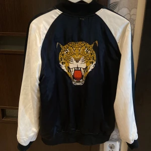 Svart och vit bomberjacka från Kendall & Kylie - En svart och vit bomberjacka från Kendall & Kylie med ett broderat tigerhuvud på ryggen och texten 'Wild at Heart' på framsidan. Jackan har långa ärmar och dragkedja framtill.