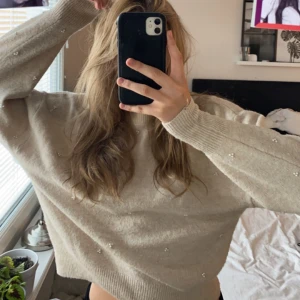Beige stickad tröja med pärlor - En beige stickad tröja med pärlor. Tröjan har långa ärmar och ribbstickade muddar vid ärmslut och nederkant. Den har en hög krage och är dekorerad med små pärlor.