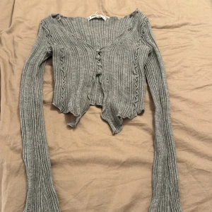 Grå ribbad kofta - En grå ribbad kofta med långa ärmar och knappar framtill. Koftan har en vågig kant vid ärmslut och nederkant, vilket ger den en unik och stilfull look.