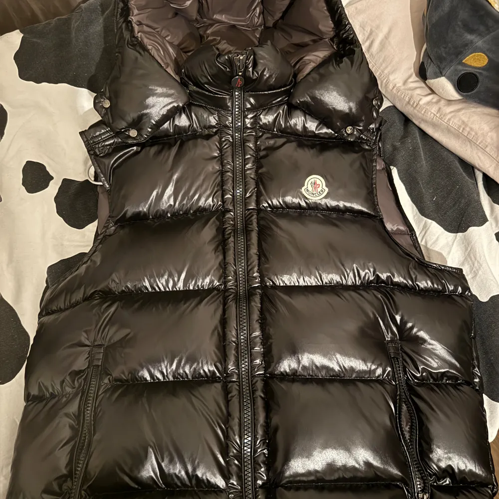 En svart, glansig dunjacka från Moncler med dragkedja framtill. Jackan har en quiltad design och är ärmlös, vilket gör den perfekt för lager-på-lager under kallare dagar. Passar dig som är mellan 170-185cm. Jackor.