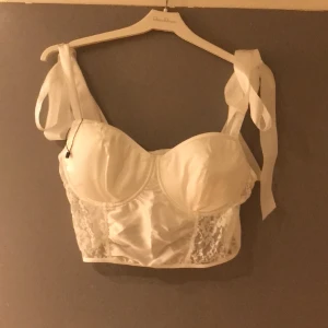 Bustier/korsettliknande oanvänd str L - Oanvänd i vit mjuk satäng med Knut axlar sidenband. Hyskknäppning bak med två olika bredd ryggen. Vaddering byst samt båge som formar. Stretchigt 90C-85D. Går ha som den är under kavaj eller som underplagg. Går även ha som burlesk klädsel. 
